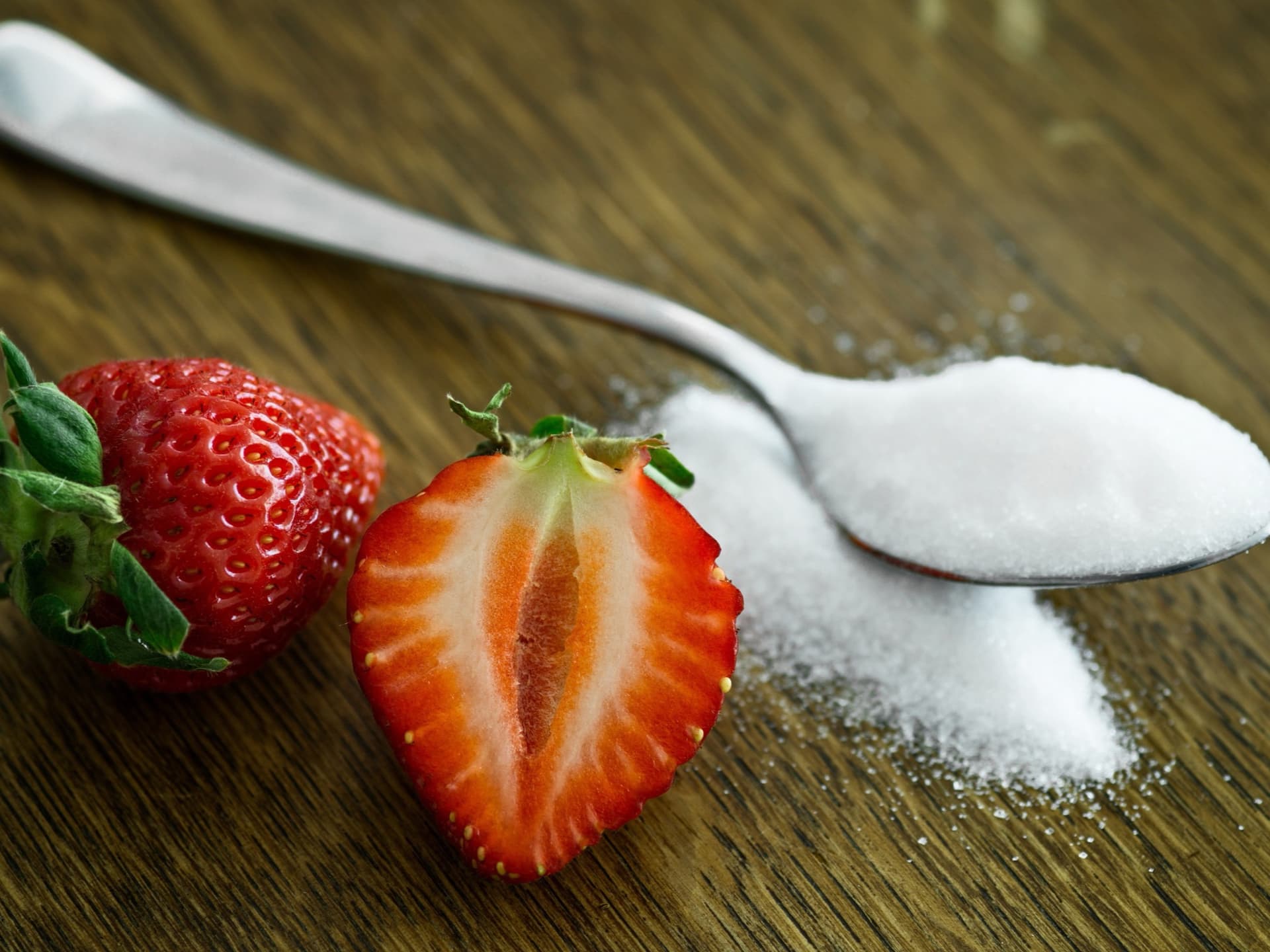 Halved strawberry beside a spoonful of white sugar on a wooden table
